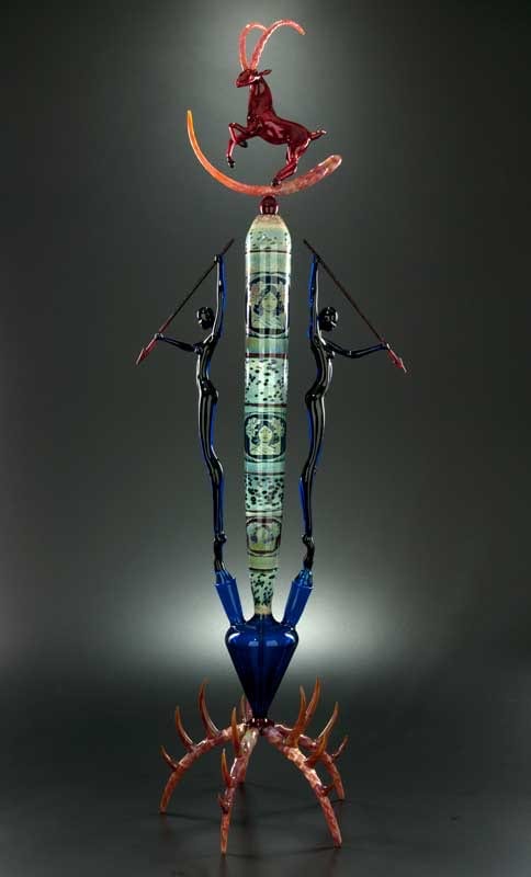A 42-inch flameworked glass sculpture by Robert Mickelsen: a tall column patterned in blue and green, held up by two slender figures, with a red ibex-like creature leaping at the top and a red crab-like form at the base