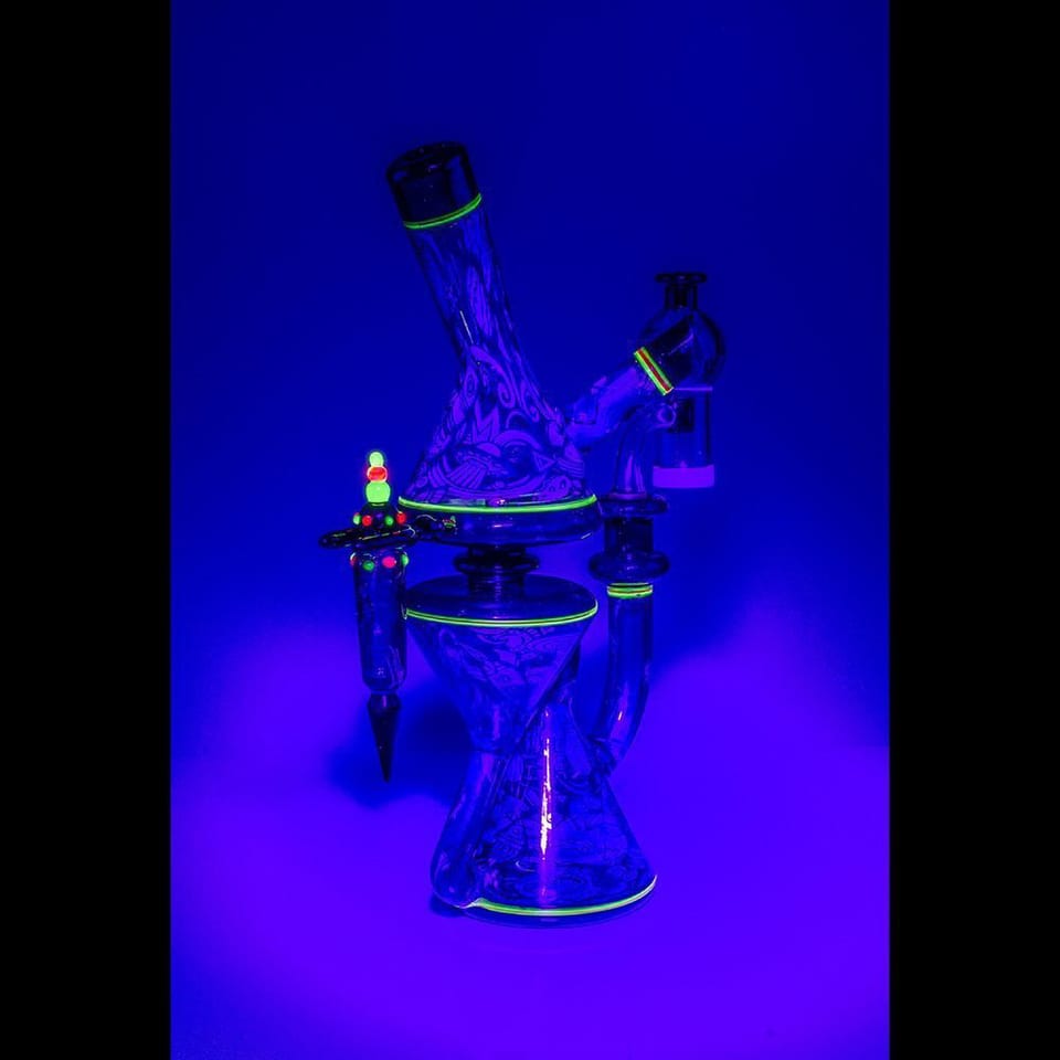 Glass recycler by Robert Mickelsen photographed under ultraviolet light, glowing blue with green accents on the rings and joint
